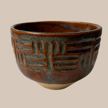 A handmade ceramic cup with a rustic, hand-painted finish. The bowl features a dark brown and black glaze with carved geometric patterns on the exterior, and a natural beige clay foot at the base, set against a solid beige background.