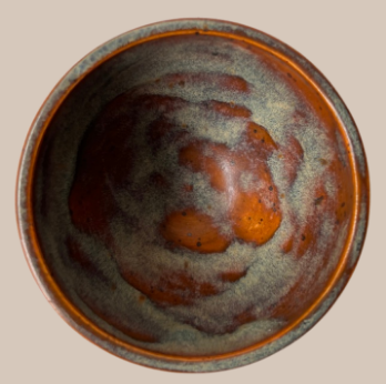 A top-down view of a round ceramic cup with a rustic, hand-painted glaze featuring mottled brown, orange, and gray colors on a light beige background.