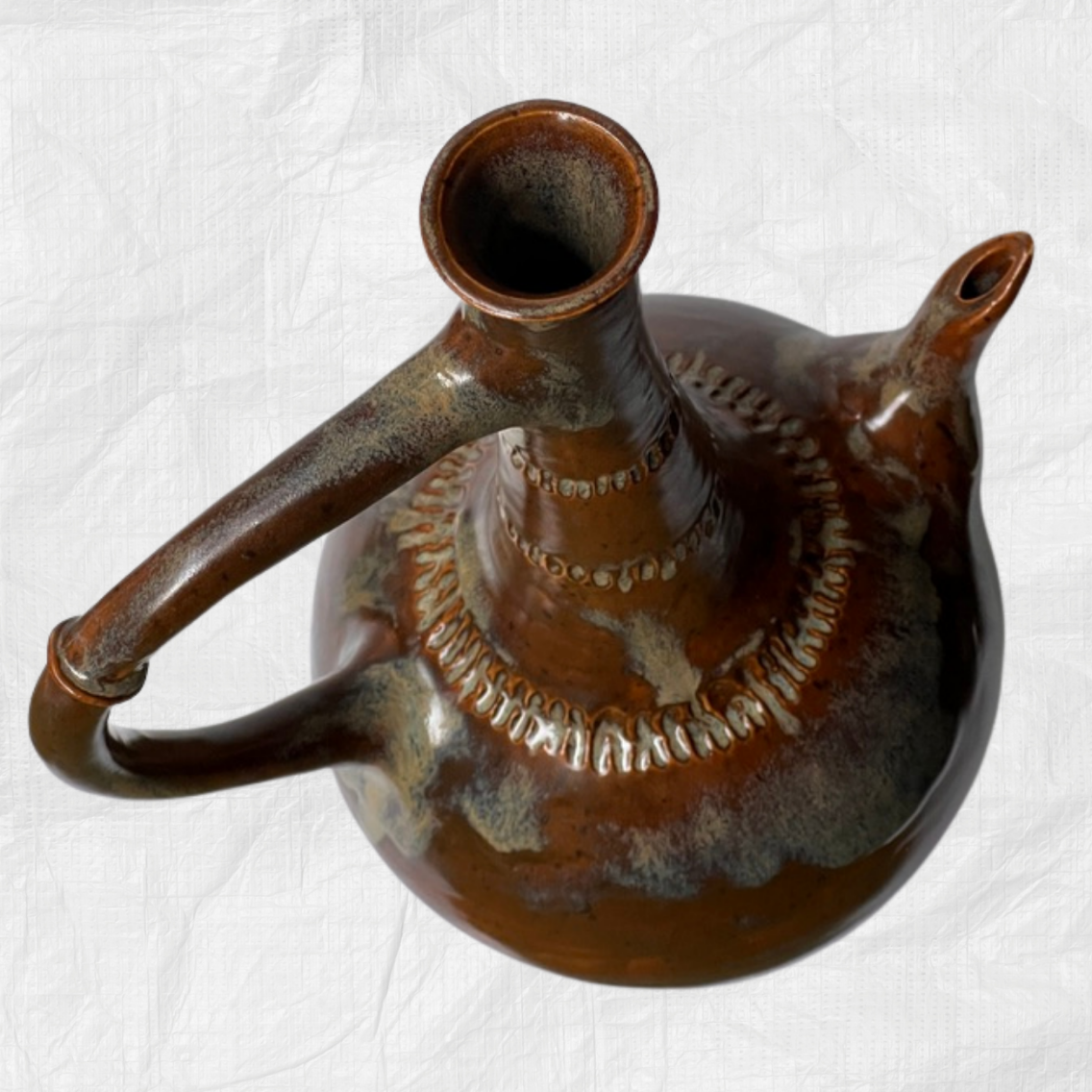 A handmade ceramic coffee pot with a rounded body, curved handle, and spout, featuring a glossy brown glaze with earthy blue-green and beige variations, set against a wrinkled white paper background.