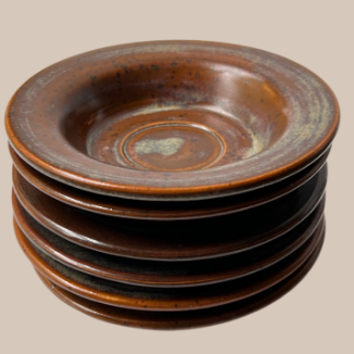 A stack of six ceramic saucers with a glossy, rustic brown glaze and subtle greenish patina accents, set against a solid beige background.